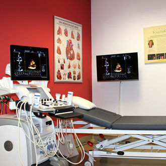 Behandlungsraum mit Liege und roter Wand mit Anschauungsbild vom menschlichen Herz in der Kardiologie