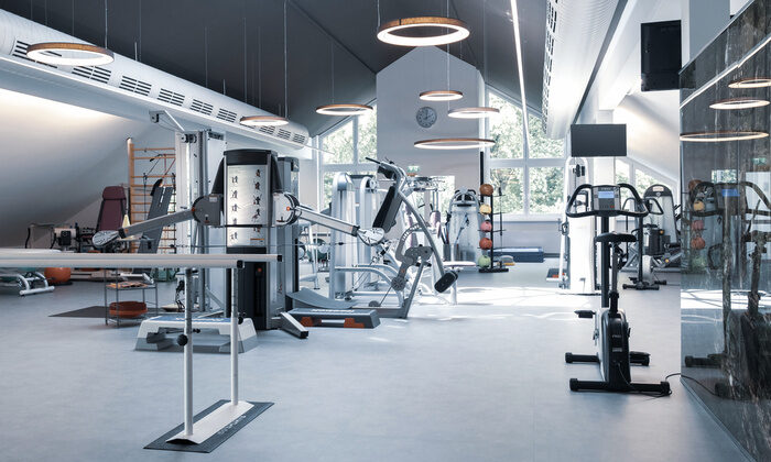 St. Wolfgang Sportpark als Fitnessstudio mit modernen Trainingsgeräten, darunter Laufband und Hanteln. Helle Beleuchtung aus runden Deckenlampen. Große Fenster bieten Blick auf grüne Bäume. Holzgeländer im Vordergrund.