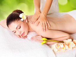 Person erhält Rückenmassage, liegt entspannt mit einer Blüte im Haar. Die Umgebung ist hell und beruhigend, mit weichen Handtüchern und dekorativen Blumen.