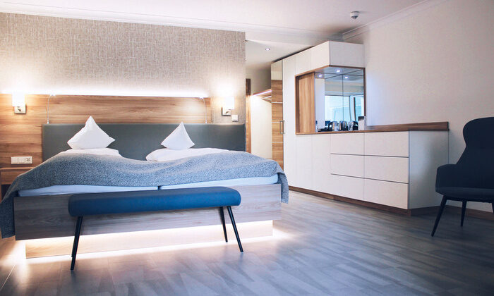 Modernes Doppelbett mit grauer Decke und weißen Kissen steht in einem stilvollen Hotelzimmer. Rechts befinden sich weiße Schränke und ein gepolsterter Stuhl. Ambiente ist elegant und einladend.