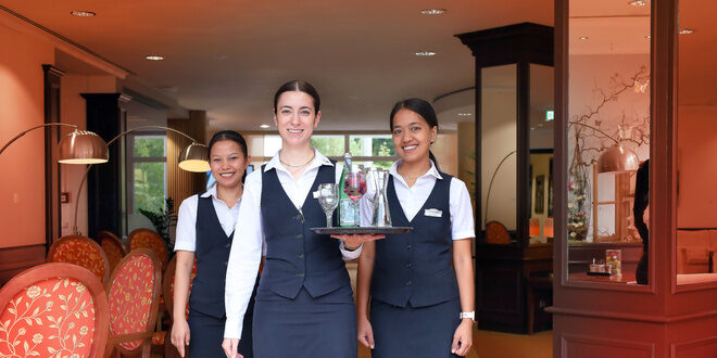 Three smiling hotel staff members stand together, with one carrying a tray of glasses and a bottle, in an elegant, warmly-lit dining area with red chairs and modern decor.