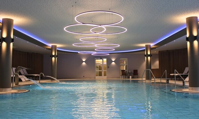 Thermal-Innenpool mit Liegen umgeben, moderne Lichter an der Decke, Holzwandverkleidung, große Fenster, entspannte Atmosphäre, in Klinik und Hotel St. Wolfgang.