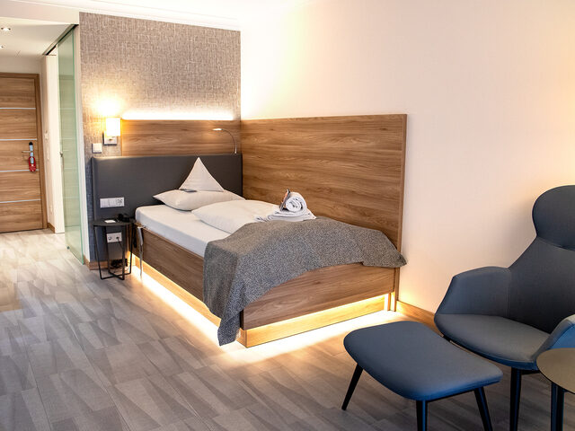 A modern room features a single bed with a bedside control panel, adjacent to two chairs and a small table.