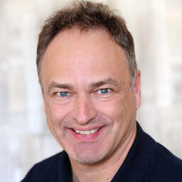 Prof. Dr. Angele, Orthopäde vom sporthopaedicum (Spezialgebiet Knie, Fraktur- und Knorpelbehandlung), steht lächelnd im Vordergrund. Er trägt ein dunkles Poloshirt. Der Hintergrund ist unscharf und hell, was eine freundliche Atmosphäre vermittelt.