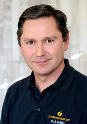 Dr. Schütz, Orthopäde (Spezialgebiet Hand), trägt ein dunkles Poloshirt mit einem Logo, schaut direkt in die Kamera, vor einem hellen, unscharfen Hintergrund. Text auf dem Shirt: „SPORTHOPAEDICUM“.