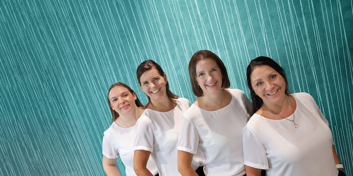 Five women stand in a diagonal line, all wearing white shirts and smiling, against a teal background with vertical lines.