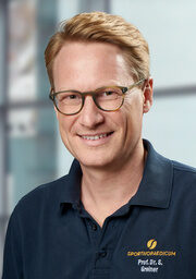 Prof. Dr. Greiner, Orthopäde (Spezialgebiet Schulter- und Ellenbogengelenk), lächelt und trägt eine Brille sowie ein dunkelblaues Poloshirt mit der Aufschrift „SPORTHOPAEDICUM“, vor einem verschwommenen Hintergrund in einem modernen Innenraum.
