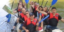 Eine Gruppe von Menschen sitzt in einem Schlauchboot und paddelt auf einem Fluss. Alle tragen rote Schwimmwesten und sind entspannt und fröhlich. Die Umgebung ist von Wasser umgeben.