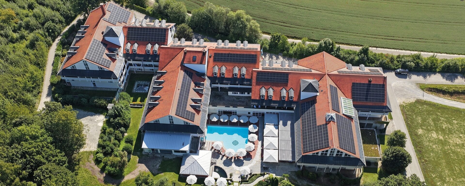 Blick aus der Luft auf Klinik und Hotel St. Wolfgang. Das große Gebäude mit roten Dächern umgibt einen Süßwasseraußenpool mit Sonnenschirmen. Es liegt neben einem Feld, eingebettet in eine grüne Gartenlandschaft. Drohnenansicht des Hotels.