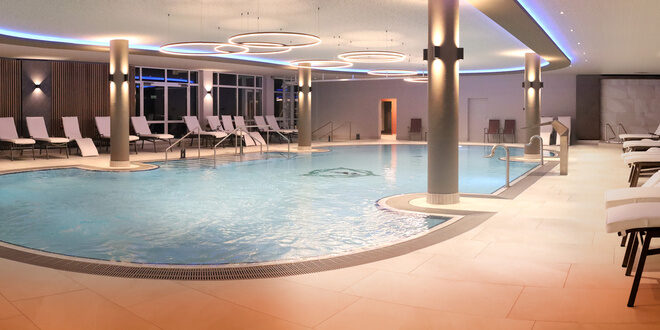 Der St. Wolfgang Thermalinnenpool mit klarem, blauem Wasser in einem modern beleuchteten Raum. Zahlreiche Liegen umgeben den Pool, unterstützt von eleganten Säulen. Die stilvolle Beleuchtung erzeugt eine entspannende Atmosphäre.