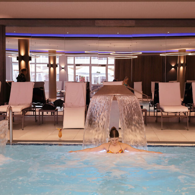 ine Frau entspannt in einem hellen Thermalinnenpool unter einem Wasserfall, umgeben von weißen Liegestühlen in einem modernen, eleganten Innenbereich.
