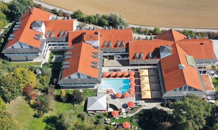 Blick aus der Luft auf Klinik und Hotel St. Wolfgang. Das große Gebäude mit roten Dächern umgibt einen Süßwasseraußenpool mit Sonnenschirmen. Es liegt neben einem Feld, eingebettet in eine grüne Gartenlandschaft.